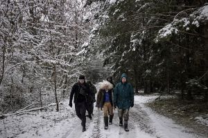 Kilka osób idaca lesnądrogą w zimowej scenerii. Ośnieżone drzewa iglaste i widoczny padający śnieg.