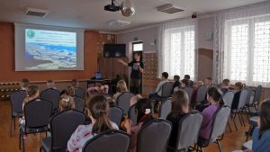 Sala, widok od tyłu. W pierwszym planie siedzące dzieci na krzesłach, w drugim planie prelegent i ekran ze zdjęciem...