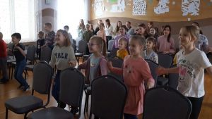 Sala wypełniona kilkudziesięciorgiem dzieci, które wykonują ćwiczenia ruchowe.