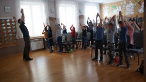 Sala wypełniona dziećmi, które wraz z prowadzącym zajęcia wykonują ćwiczenie naśladujące maskującego się w trzcinach bąka...