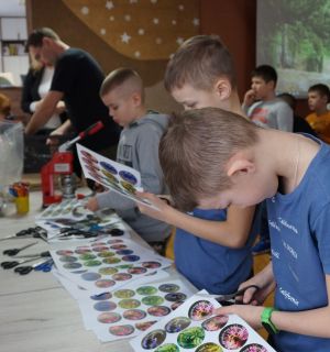 Kilkoro dzieci wycina okrągłe zdjęcia przyrodnicze. W tle, prowadzący zajęcia oprawia wycięte obrazy w metalowe przypinki.