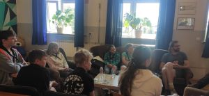 Młodzi i dorośli uczestnicy warsztatów na spotkaniu wewnątrz budynku. Siedzą w okręgu i są wyraźnie zasłuchani.