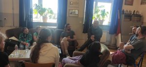 Młodzi i dorośli uczestnicy warsztatów na spotkaniu wewnątrz budynku. Siedzą w okręgu i są wyraźnie zasłuchani.