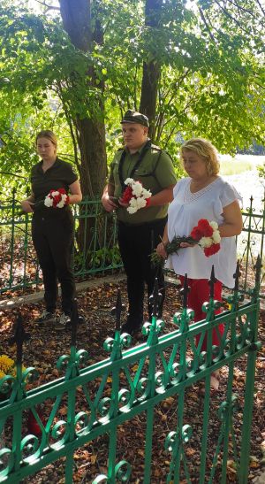 Trzy osoby stojące przy pomniku z kwiatami w rękach. Moment składania kwiatów na mogile ofiar pacyfikacji wsi Grzędy.