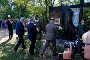Kilku mężczyzn składa kwiaty przy pomniku mieszkańców wsi Grzędy. Wkoło wielu widzów i straż żołnierska.