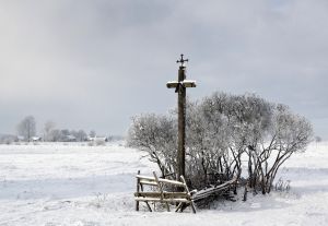 Ośnieżone pola, w pierwszym planie przydrożny krzyż, a w tle wieś.